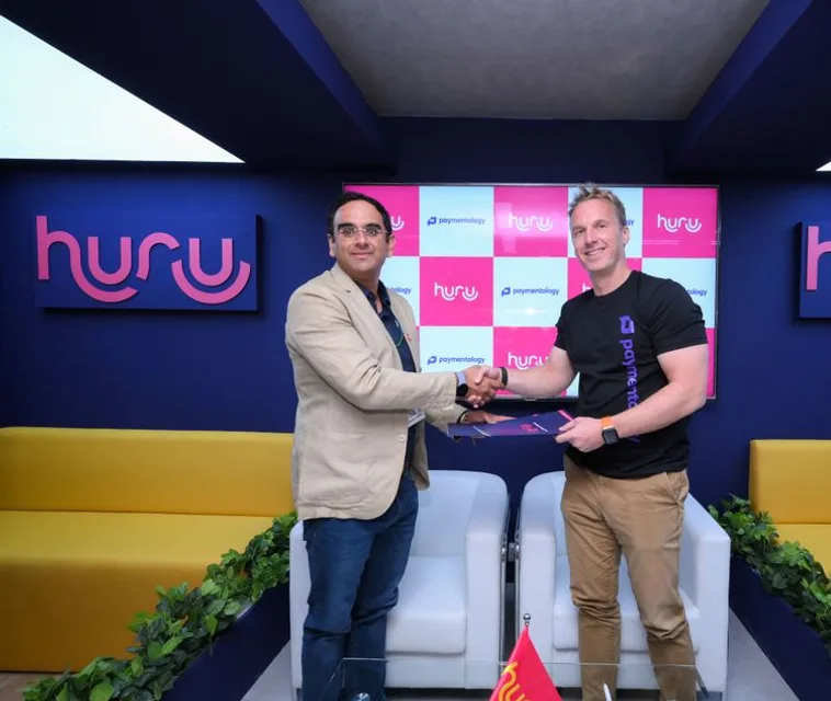 Huru and Paymentology representatives shaking hands at their MoU signing, beside a Huru logo
