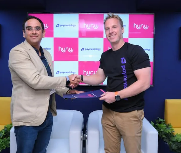 Huru and Paymentology representatives shaking hands at their partnership signing, in front of a screen with both companies’ logos